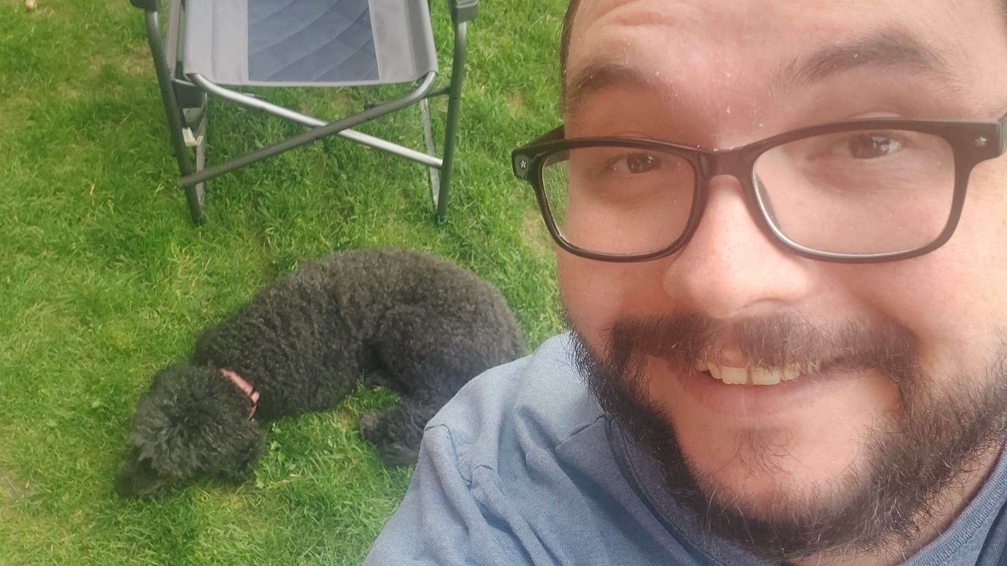 A man is dressed in a t-shirt and shorts stands next to a sleeping black standard poodle, she is sleeping and he looks stoned